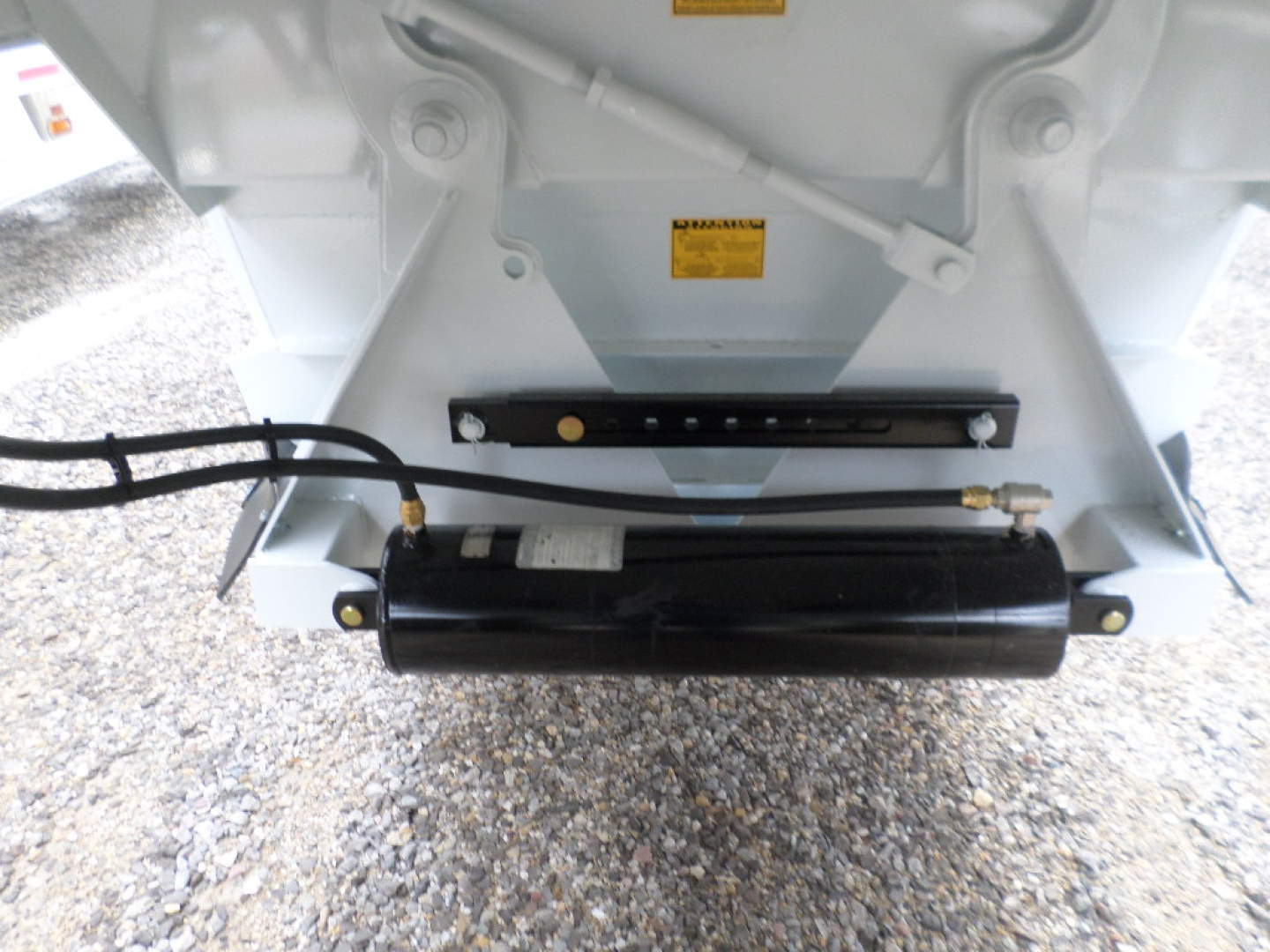 Trailer rear with air ride suspension and hydraulic component; black cylindrical actuator; warning labels on white frame; hoses connected.