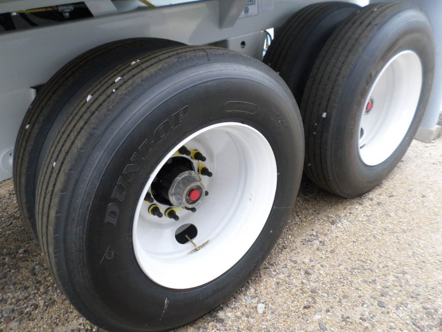Dual tandem axle trailer wheels with white rims and Dunlop tires, on a gravel lot; shows wheel hubs and suspension area.