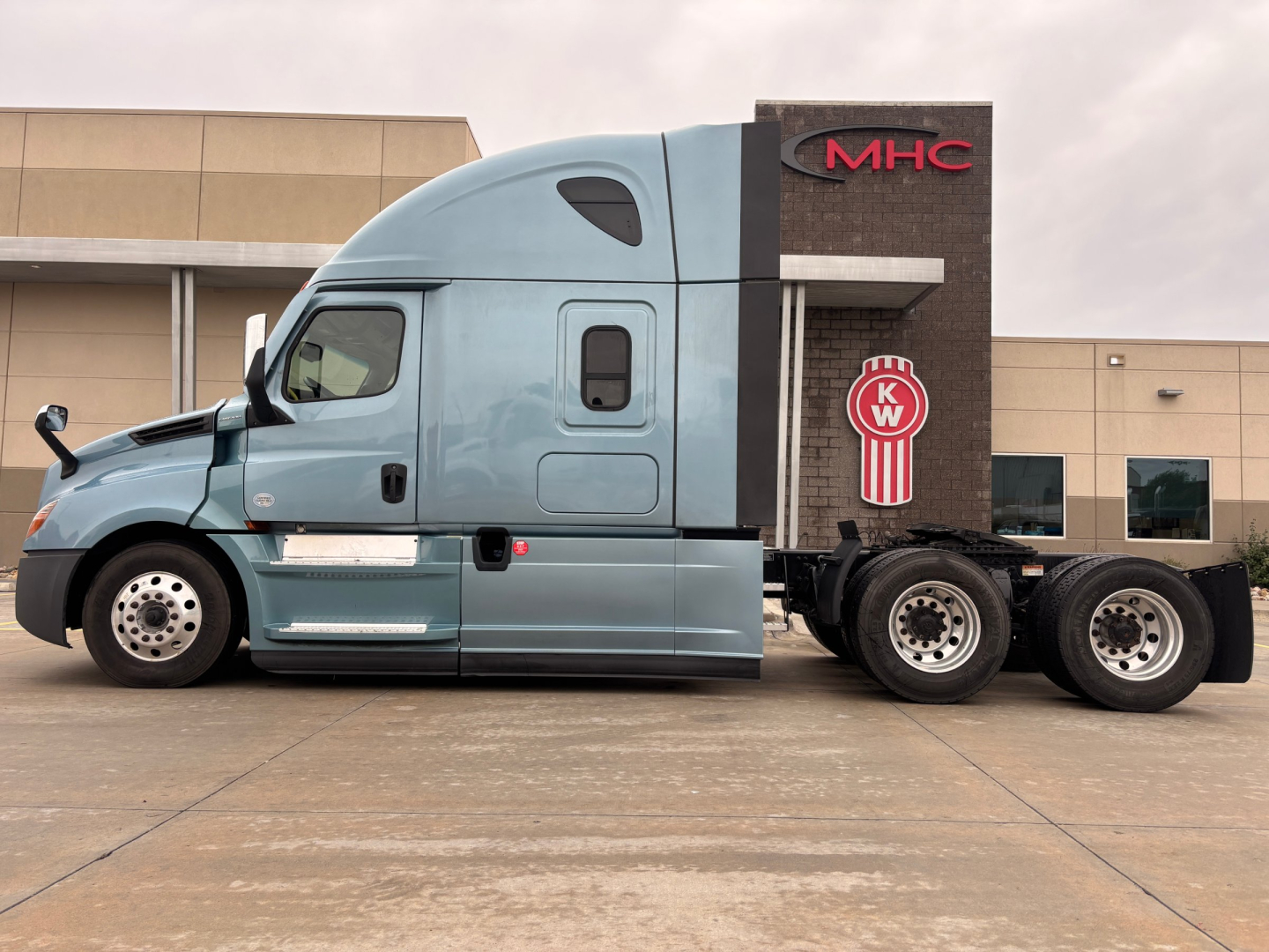 Truck side view showing a 2023 Freightliner PT126064ST with blue cab, sleeper, and tandem rear axles in an industrial lot.