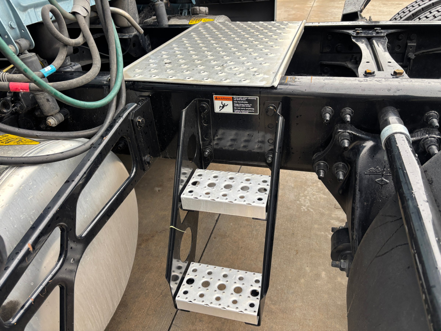 Truck steps and rear cab area showing aluminum perforated steps attached to a Freightliner chassis with hydraulic lines and a diamond-plate platform.