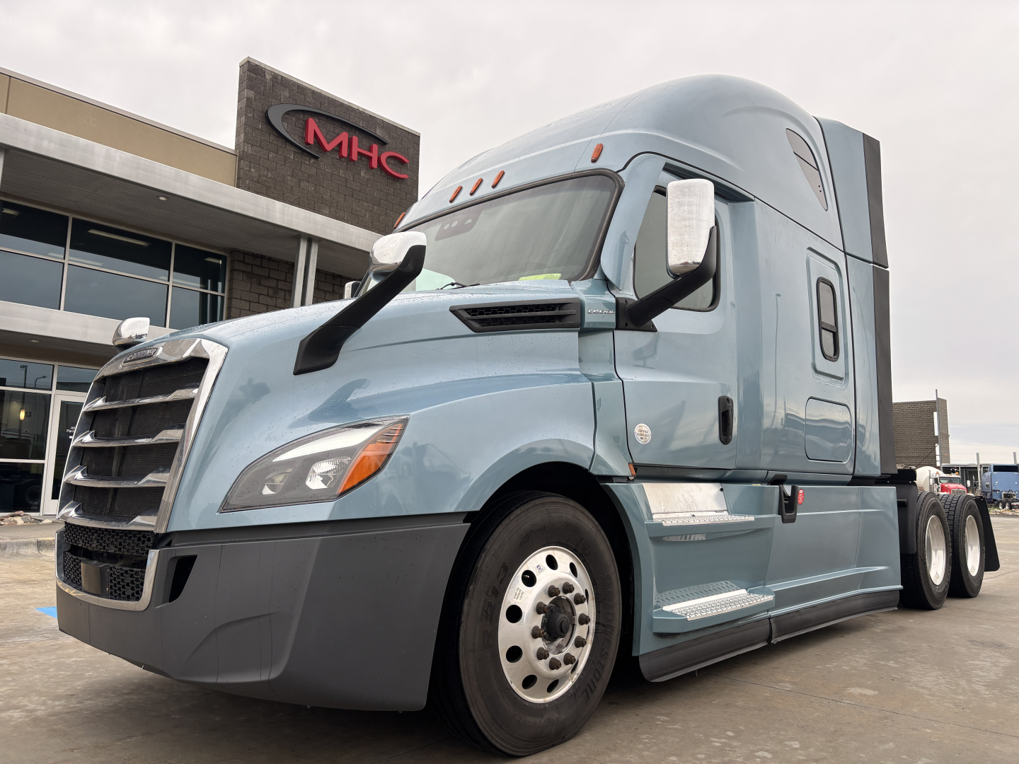 Freightliner semi truck in blue sleeper cab, side view showing badges and steps, in front of dealership building