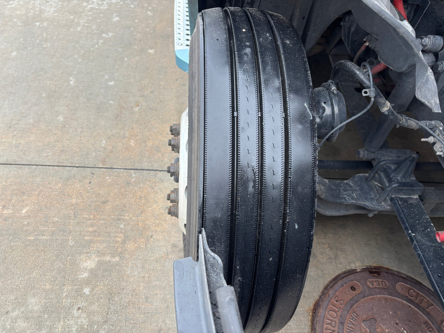 Wheel and front axle area of a Freightliner truck showing a black triple-ribbed dual rear? front drive wheel and hub with lug nuts; visible frame and brake drum. Text not legible.