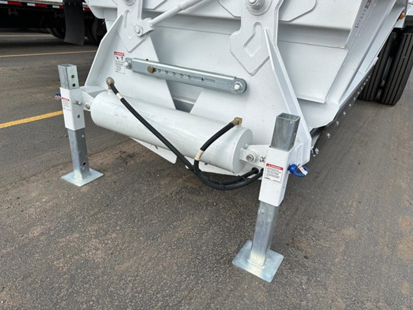 Bottom dump trailer front legs and hydraulic hoses with mounting brackets visible, white metal frame, safety pins, and support feet on concrete.