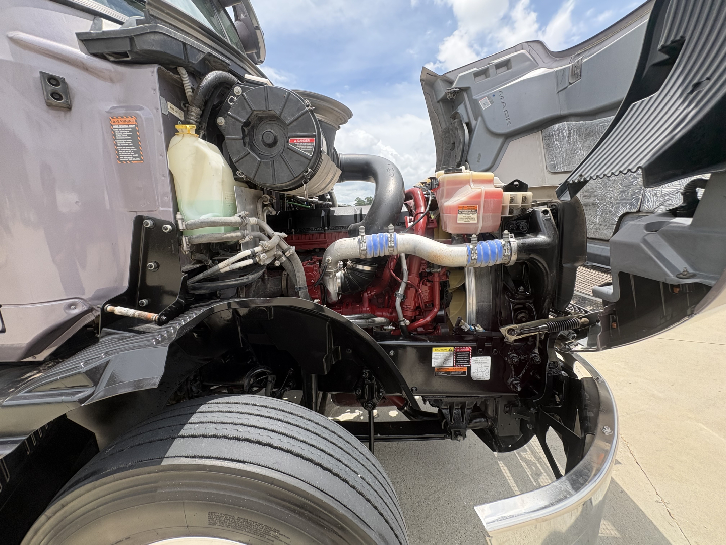 Front engine bay of a 2024 Mack AN64T70 conventional sleeper truck showing coolant reservoir, radiator hose, air intake, and engine block with visible hoses and wiring.