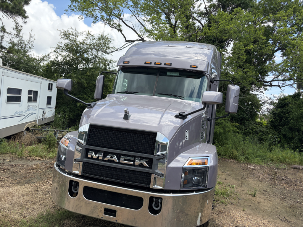 Truck front view of a gray 2024 Mack AN64T70 conventional sleeper, showing large chrome grille with MACK lettering, bumper, and dual side mirrors.