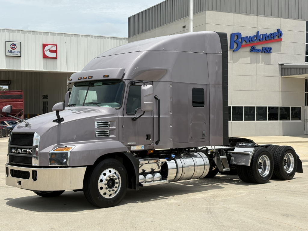 Semi truck tractor in a dealership lot showing a Mack AN64T70 with gray cab, chrome bumper, dual rear axles, fuel tanks, and Brackner's dealership building in the background.