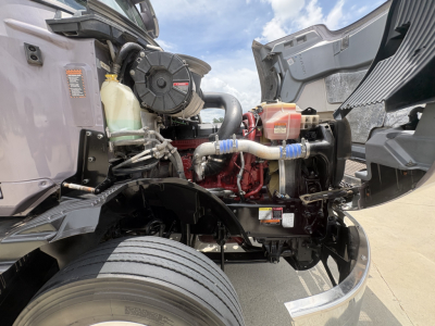 Front engine bay of a 2024 Mack AN64T70 conventional sleeper truck showing coolant reservoir, radiator hose, air intake, and engine block with visible hoses and wiring.