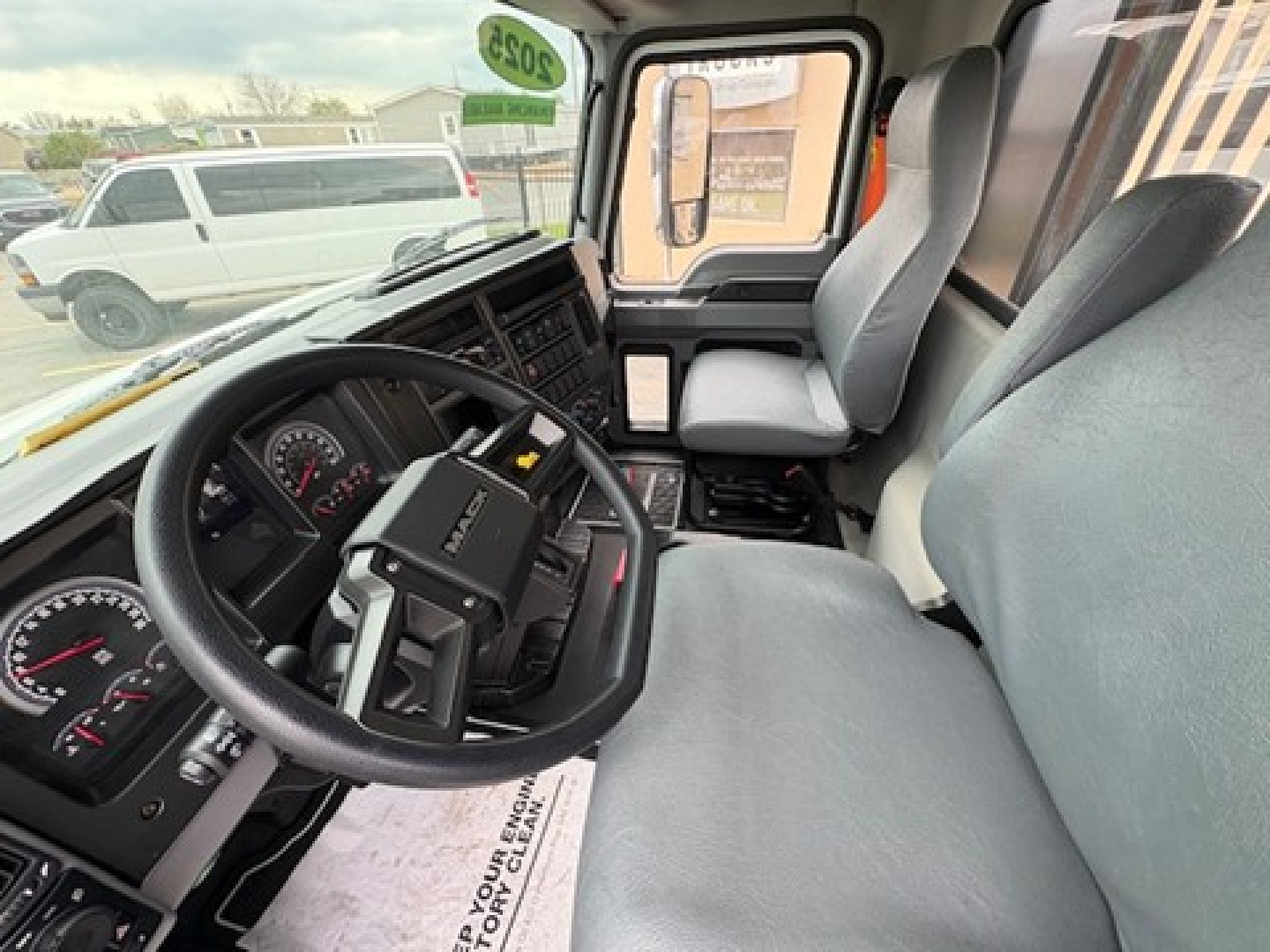 Vehicle cabin of a 2025 Mack MD6, driver seat and steering wheel in view, dashboard gauges, center console, and entry door visible.