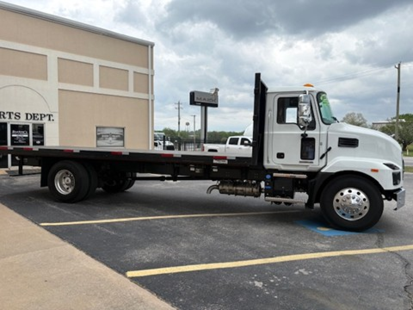 Flatbed truck with a white cab and black flatbed in a parking lot, Mack MD6 style, side view showing long flatbed and dual rear wheels.