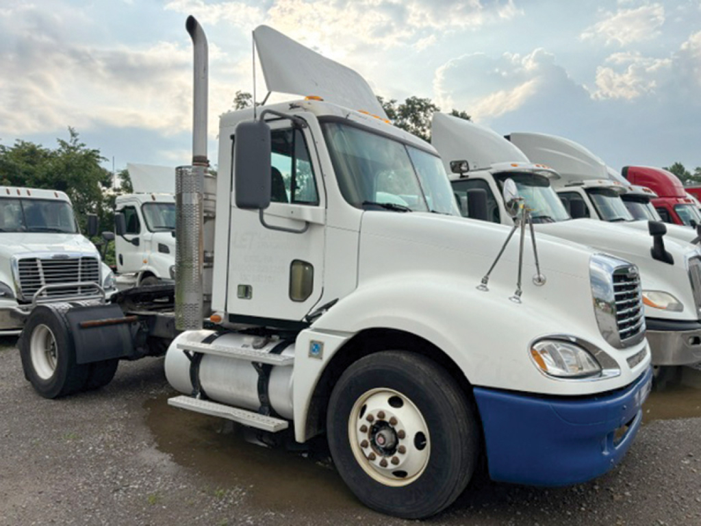 Truck cab of a white 2009 Freightliner CL120 Columbia conventional daycab with blue bumper, large exhaust stack, and parked among other tractors.