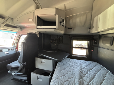 Interior cab view of a 2023 Kenworth T680 sleeper with overhead storage compartments, a retractable desk, and quilted sleeper mattress in view.