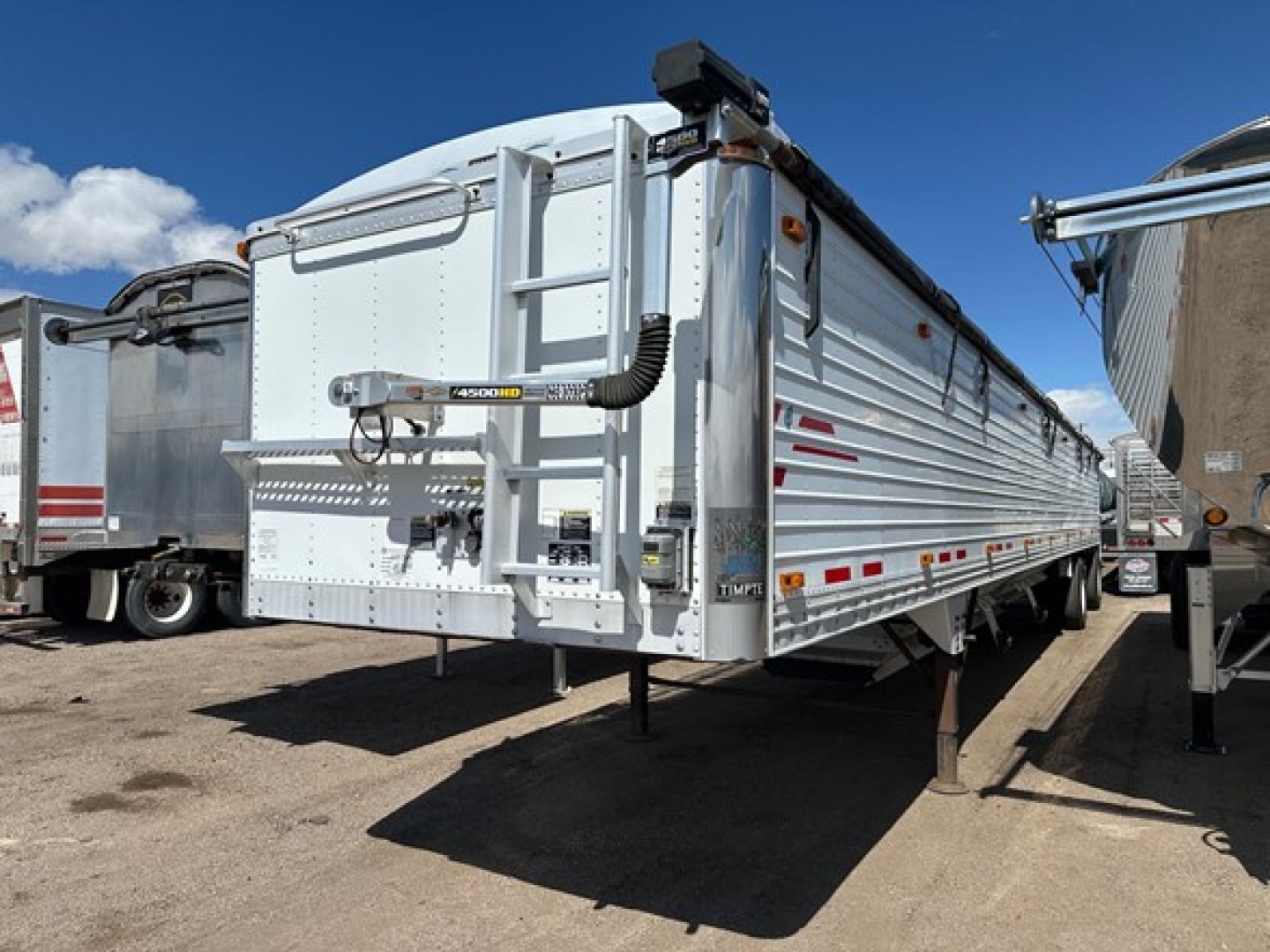 Hopper trailer in a yard showing the rear and side, a Timpte Super Hopper with silver corrugated aluminum sides, rear door, ladder, and reflector strips.