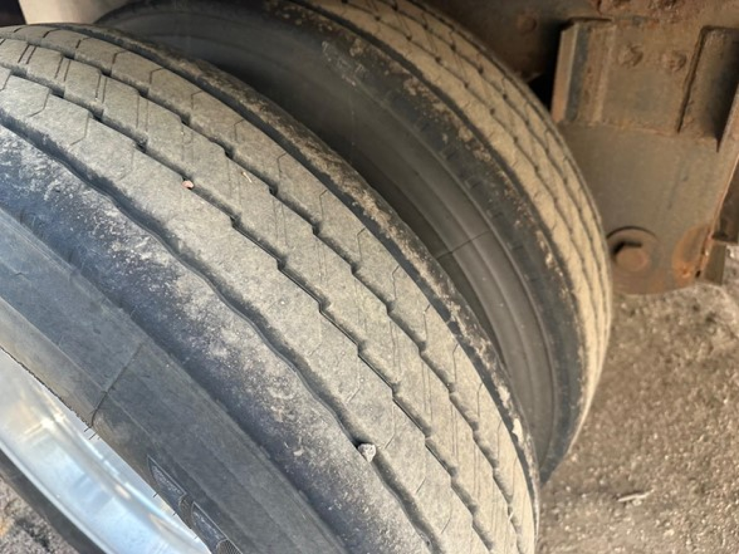 Truck trailer wheel twin tires showing worn tread and sidewall. Dirt and grime on rims and undercarriage visible.