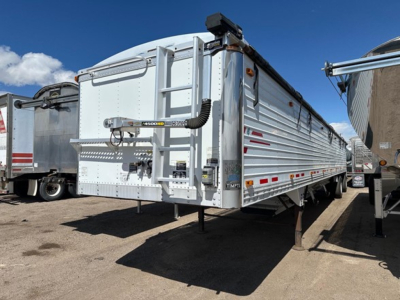 Hopper trailer in a yard showing the rear and side, a Timpte Super Hopper with silver corrugated aluminum sides, rear door, ladder, and reflector strips.