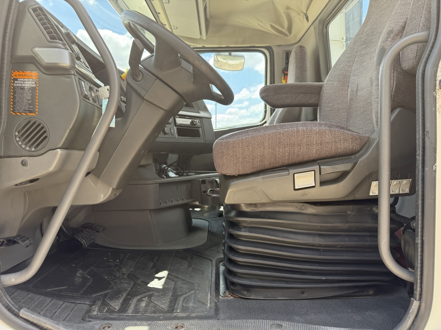 Cab interior of a Volvo VNL300 truck showing steering wheel, driver's seat, and floor mat with a beige seat cushion and ribbed base.