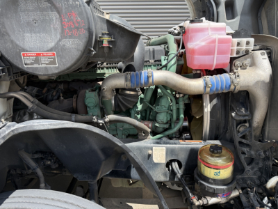 Volvo VNL300 engine bay with coolant reservoir, hoses, air filter housing, and hydraulic reservoir visible; pink coolant container, blue hose clamps, yellow-rimmed cap.