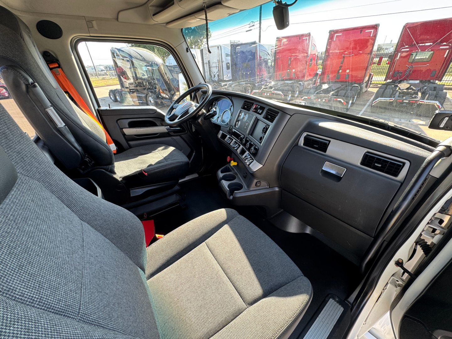 Kenworth T680 truck cabin interior with steering wheel, dashboard, and gray fabric seats. Visible pickup from driver seat showing instrument cluster and central console.