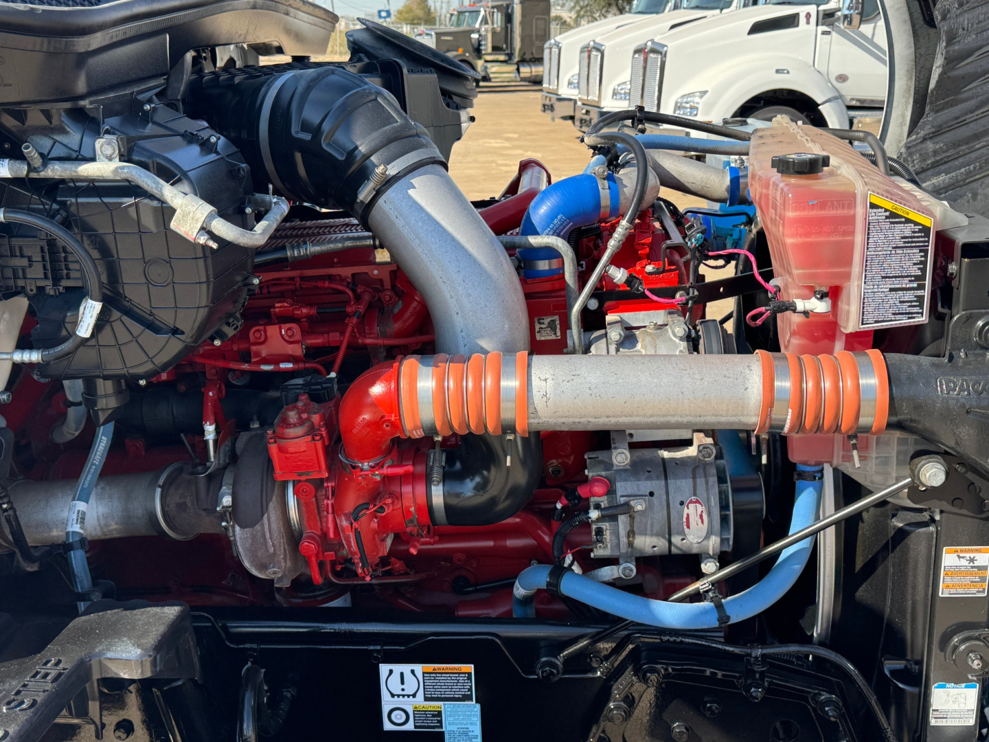Kenworth T680 engine bay showing red engine block, large air intake, orange flexible protective conduit, blue hoses, and coolant reservoir.