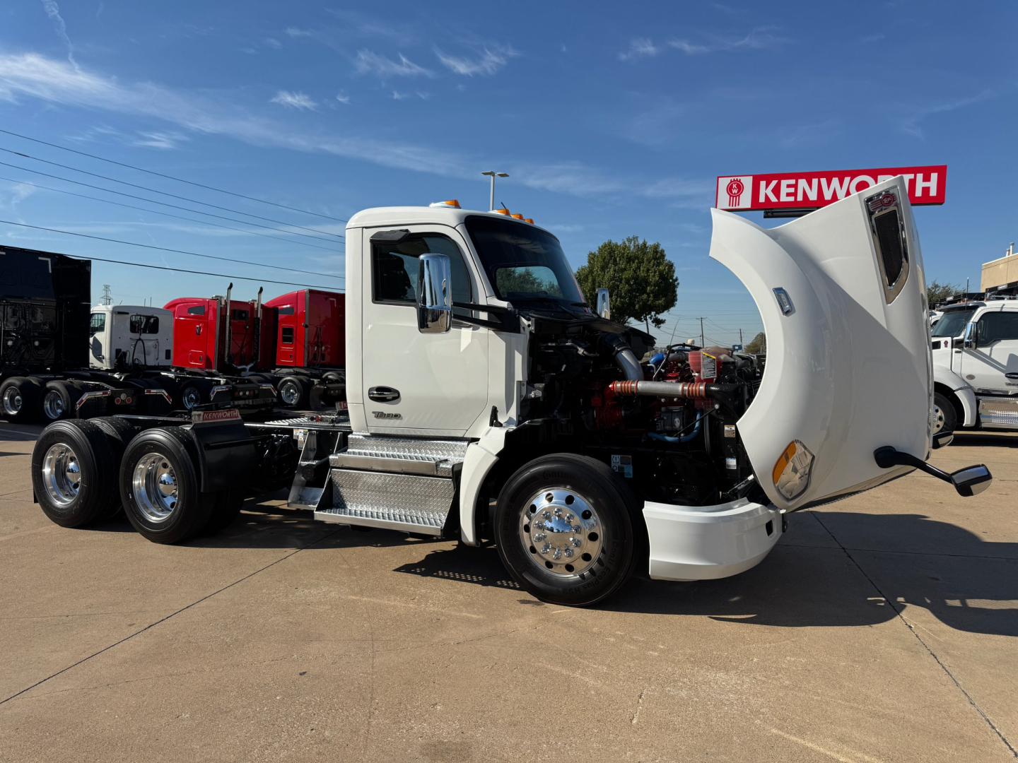 Kenworth T680 conventional daycab truck with hood open showing engine bay and cab in white, front fender, chrome wheels, and Kenworth dealership in background.