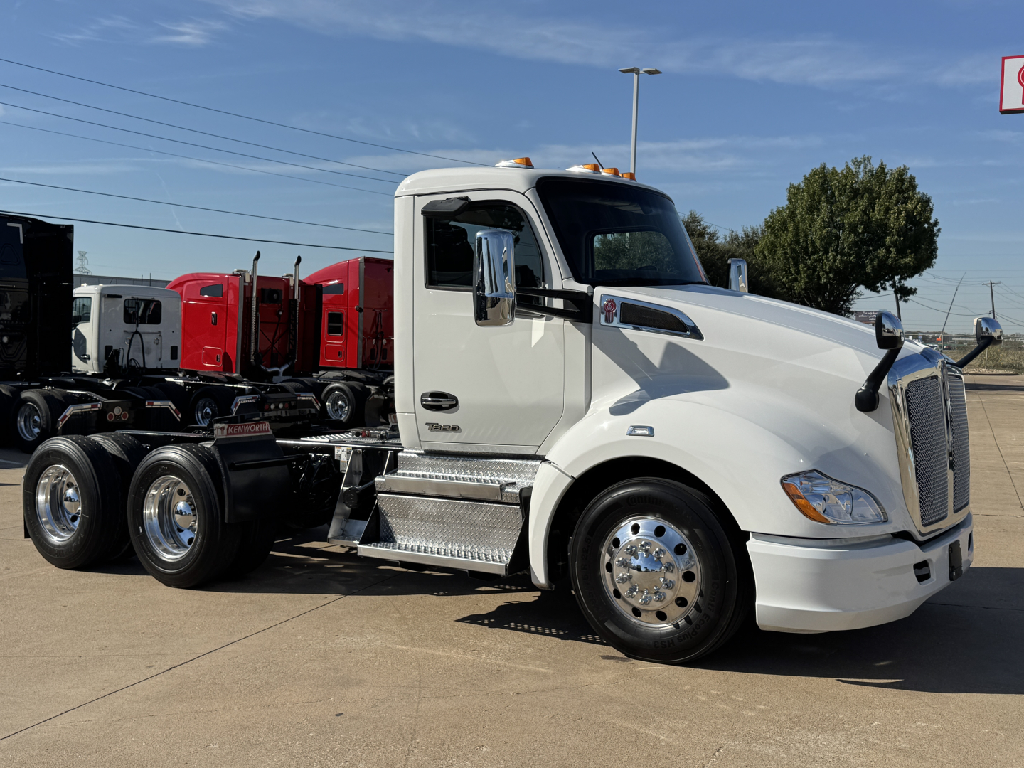 Kenworth T680 truck tractor in white, side view, with chrome fuel tanks and steps; text on truck includes 'Kenworth' emblem and side mirror fins; background shows other trucks.