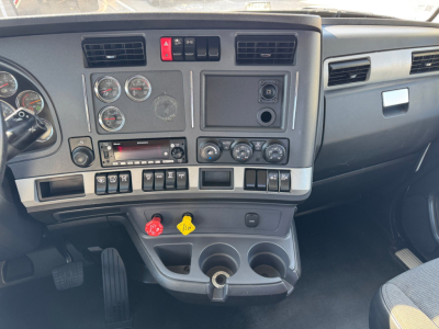 Kenworth T680 dash panel with multiple gauges, controls, vents, and a yellow handle; center stack shows climate controls, audio, and switch panel; lower console with cup holders and 12V outlet.