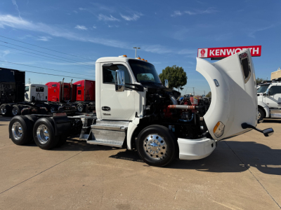 Kenworth T680 conventional daycab truck with hood open showing engine bay and cab in white, front fender, chrome wheels, and Kenworth dealership in background.