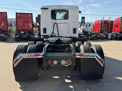Truck tractor rear view showing dual rear axles and fifth wheel mounting on a white cab with black chassis; multiple red trucks in background at lot.