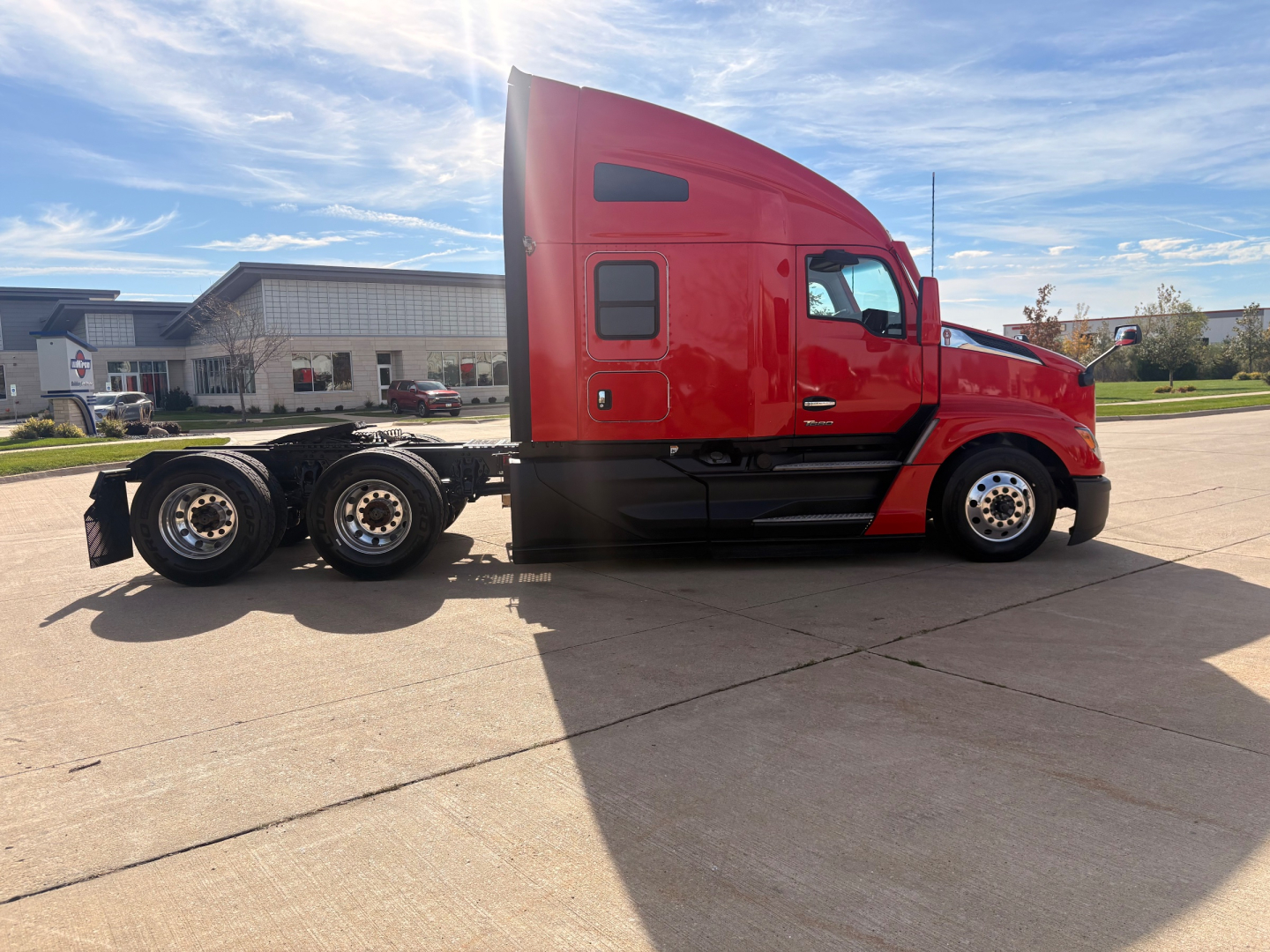 Kenworth T680 tractor side view in red, with sleeper cab and multiple wheels showing. No visible text readable on the side panels, no specific fluid labels.