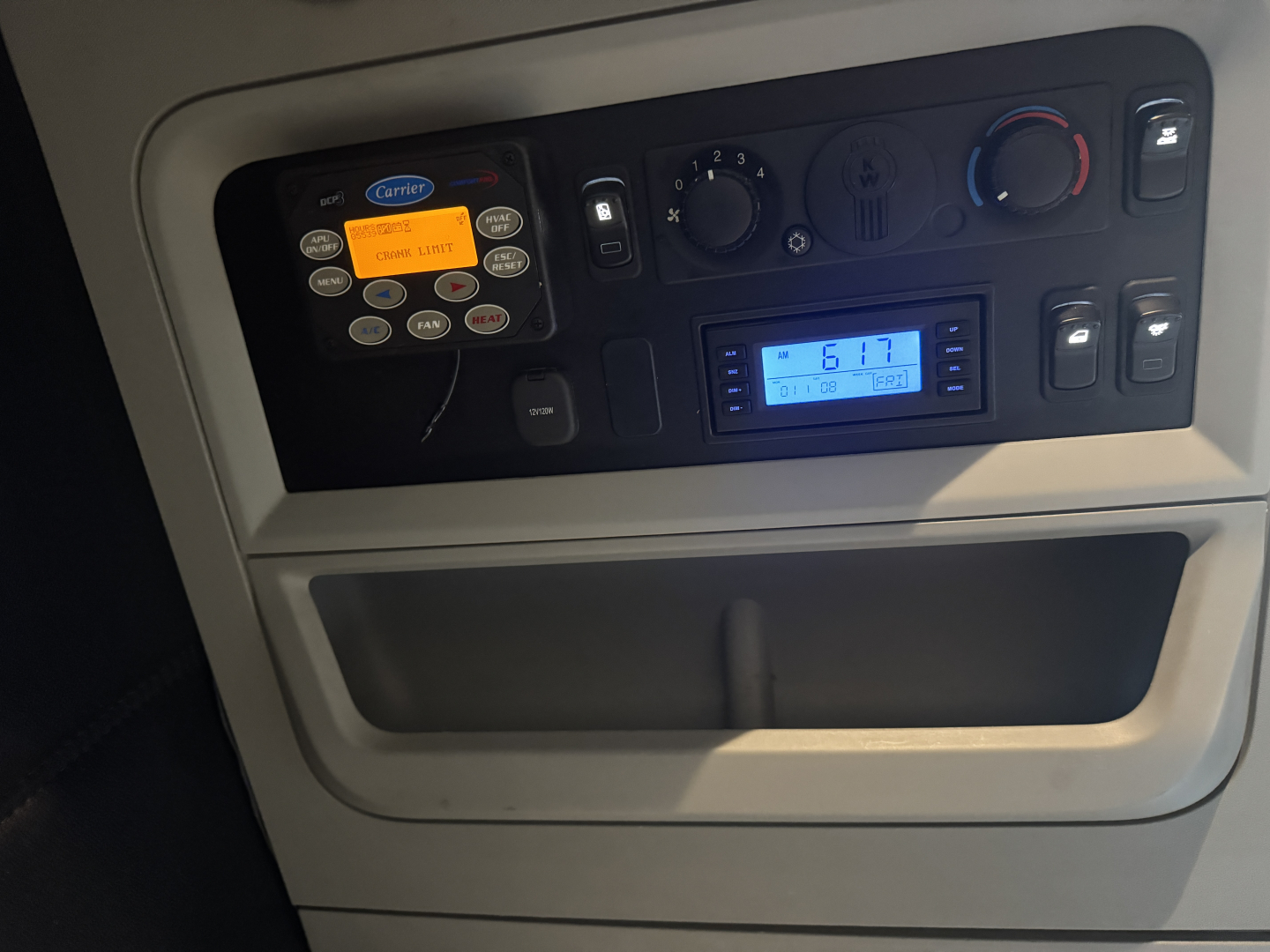 Kenworth T680 interior panel showing carrier transceiver with buttons, digital display, and climate controls. Visible text includes Carrier COMMUNICATIONS label and various control labels.