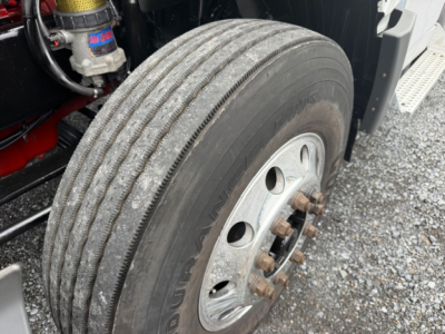 Truck wheel and tire close-up showing dual rear axle and lug nuts on a 2018 International LT625 conventional cab. Visible components include tire tread, wheel hub with rusted lug bolts, brake drum area, and part of suspension. Text on tire 