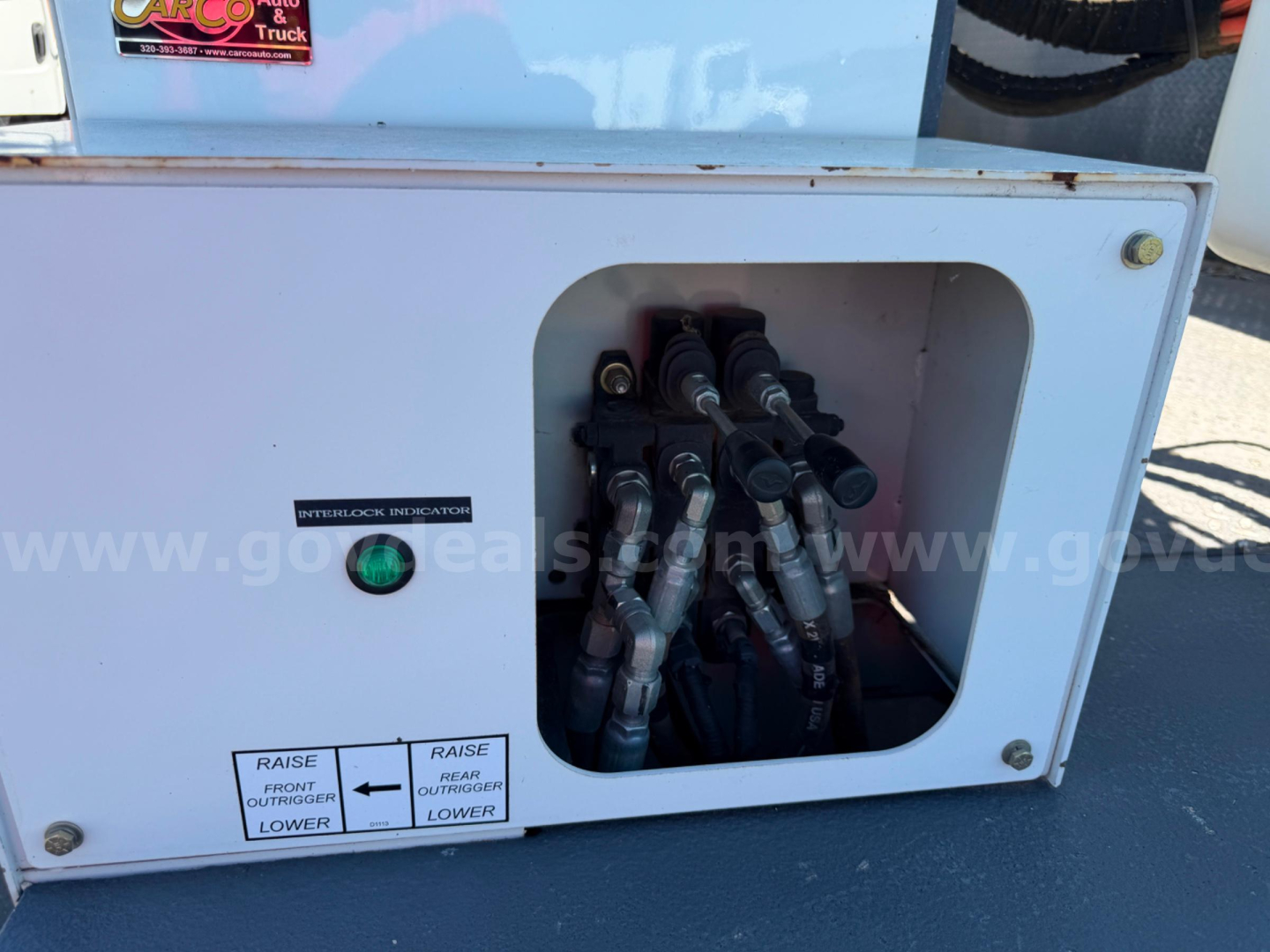 Engine bay showing multiple hydraulic/fuel lines connected to a manifold inside a metal box with a green indicator light and labeled controls.