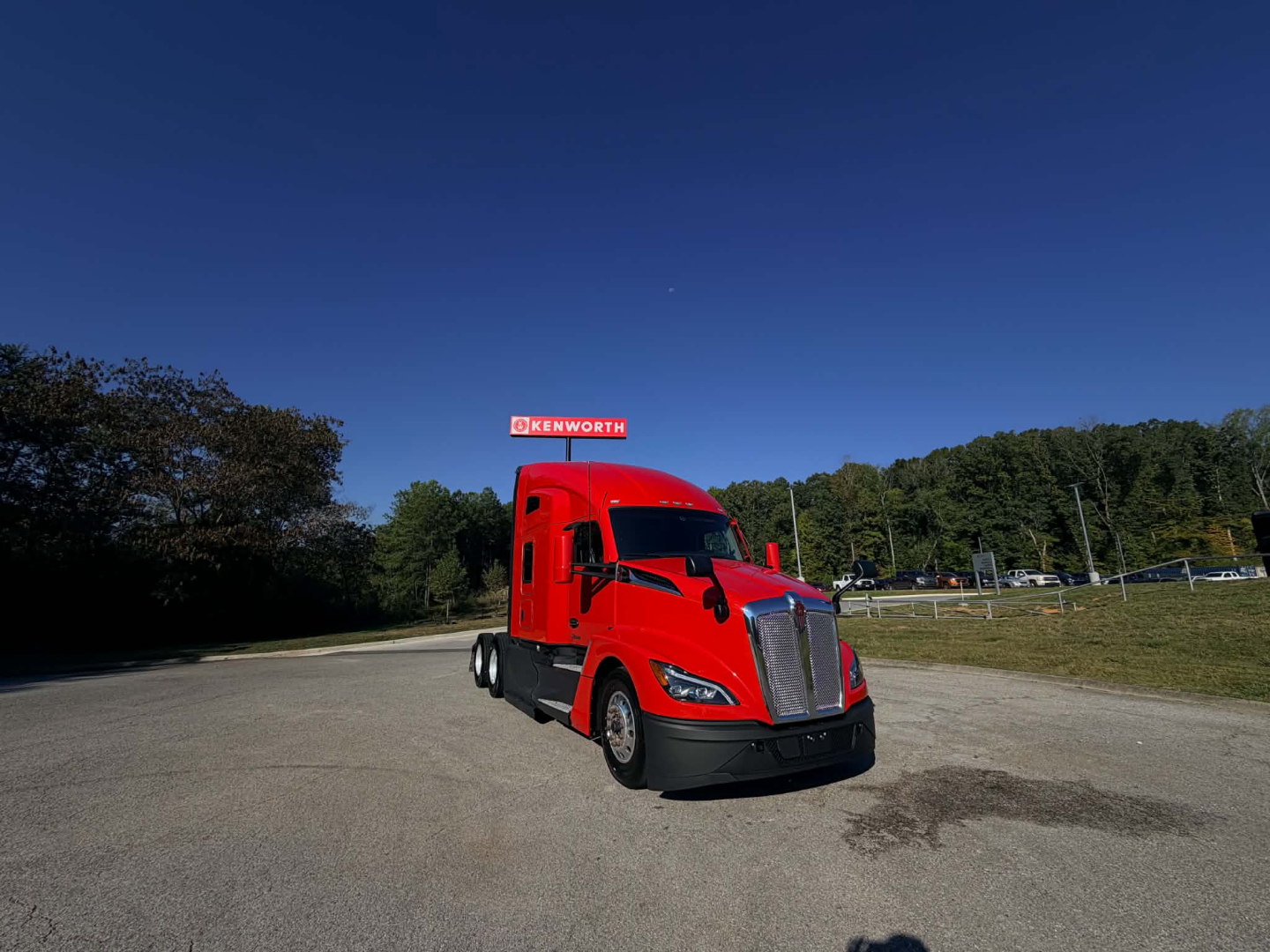 Kenworth T680 conventional sleeper truck in red, front-left view, highway lot, Kenworth dealership sign in background.