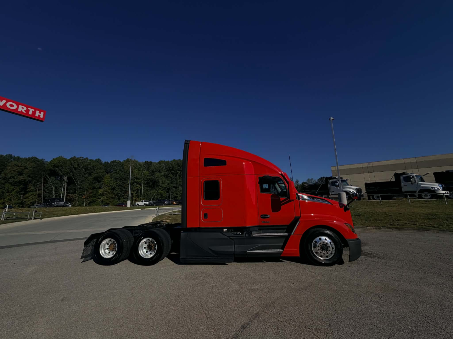 Red Kenworth T680 side view tractor in lot showing sleeper cab and triple-axle rear, clear sky.
