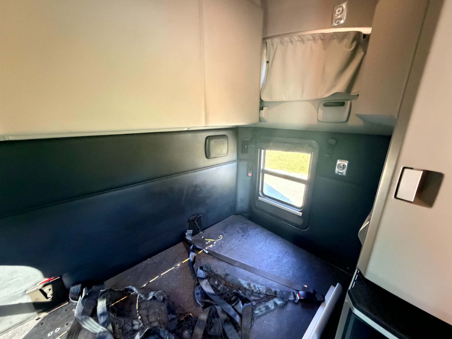 Kenworth T680 sleeper cab interior showing small side window, upper storage hatch, and seat belts on the floor in a compact space.