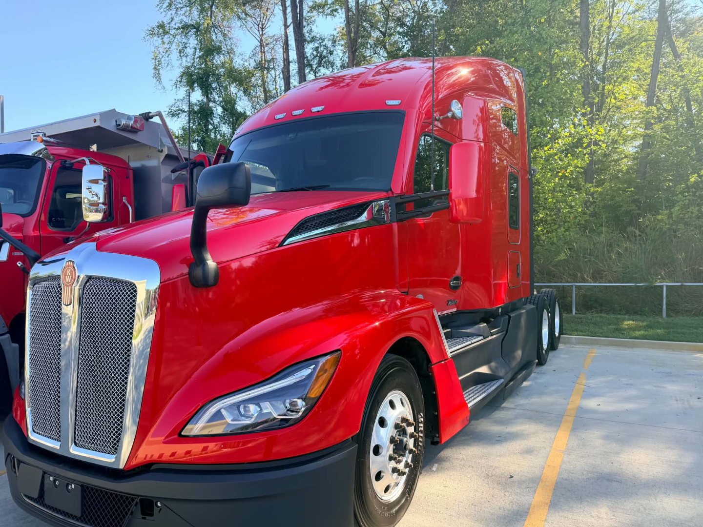 Kenworth T680 tractor in red color, front left view, showing cab and grille with chrome vertical bars, daytime outdoor lot.