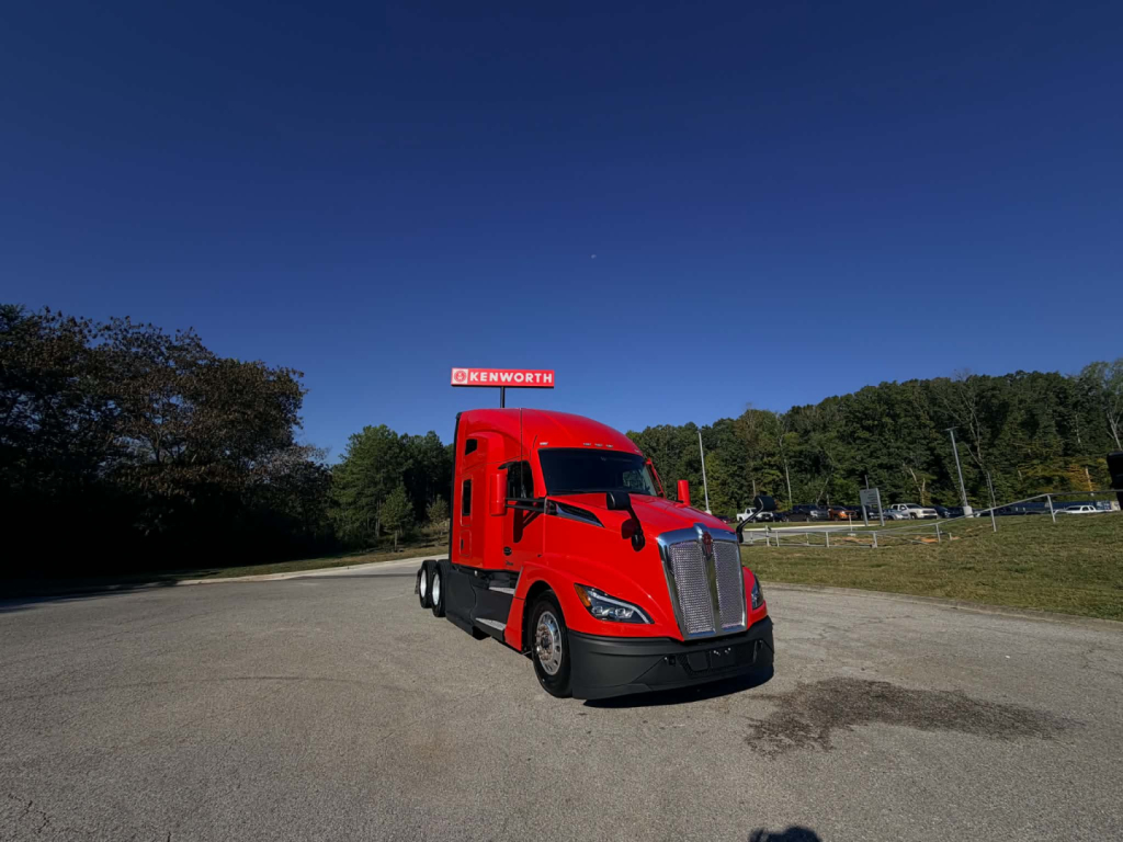 Kenworth T680 conventional sleeper truck in red, front-left view, highway lot, Kenworth dealership sign in background.