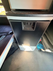 Kenworth T680 interior center console with beverage? cooler and USB/12V port panel in view. VIN and stock number not visible; image shows a black cabinet with a rectangular panel and a power outlet area.