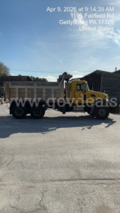 Dump truck showing yellow Mack GU713 in-service with long flatbed body and crane arm, side profile visible.