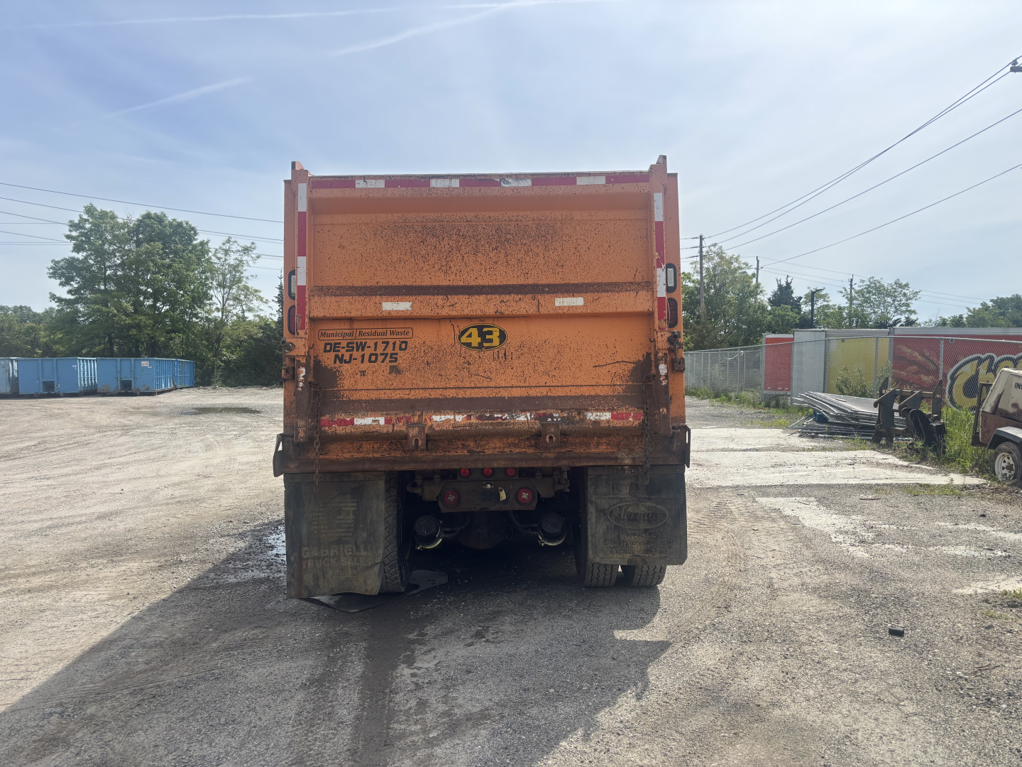 Rear view of a 2016 Kenworth T880 dump truck showing the orange dump body with mudflaps and rear hitch area. Visible text includes DE-SW-1710? NU-1075? and a yellow circular warning sticker with 43.