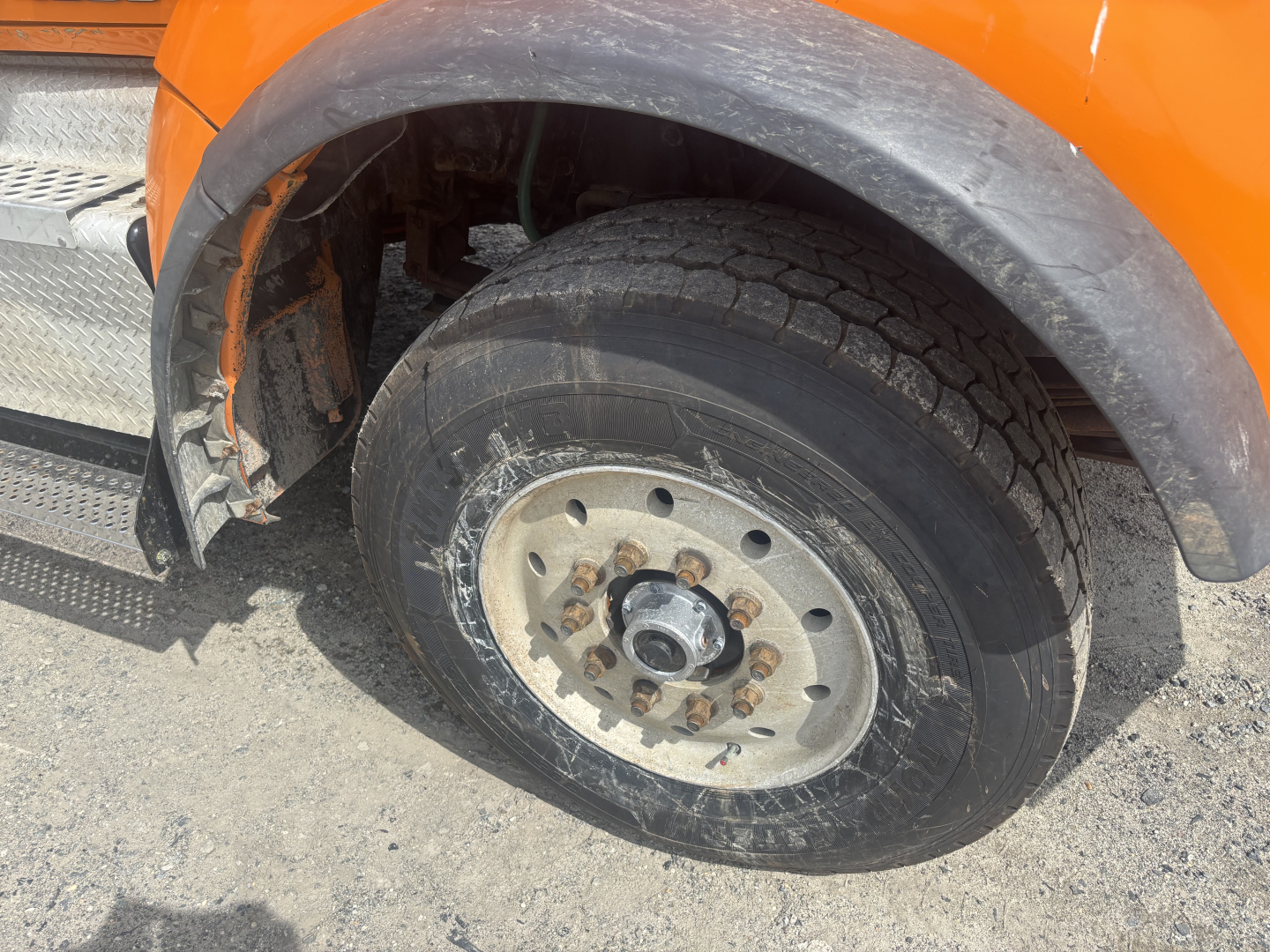 Truck front wheel area of a 2016 Kenworth T880 dump truck showing a large tire with a metal wheel, lug nuts, and wheel well; orange body panel and side step visible.