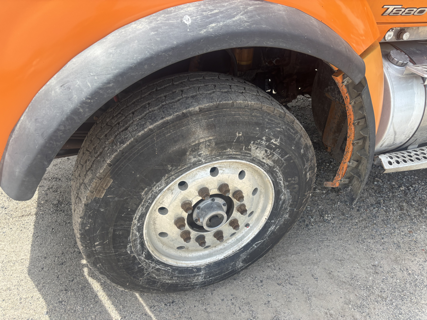Front wheel and fender of an orange 2016 Kenworth T880 dump truck showing wheel with dirt and lug nuts, partial hood and side badge 'T880'