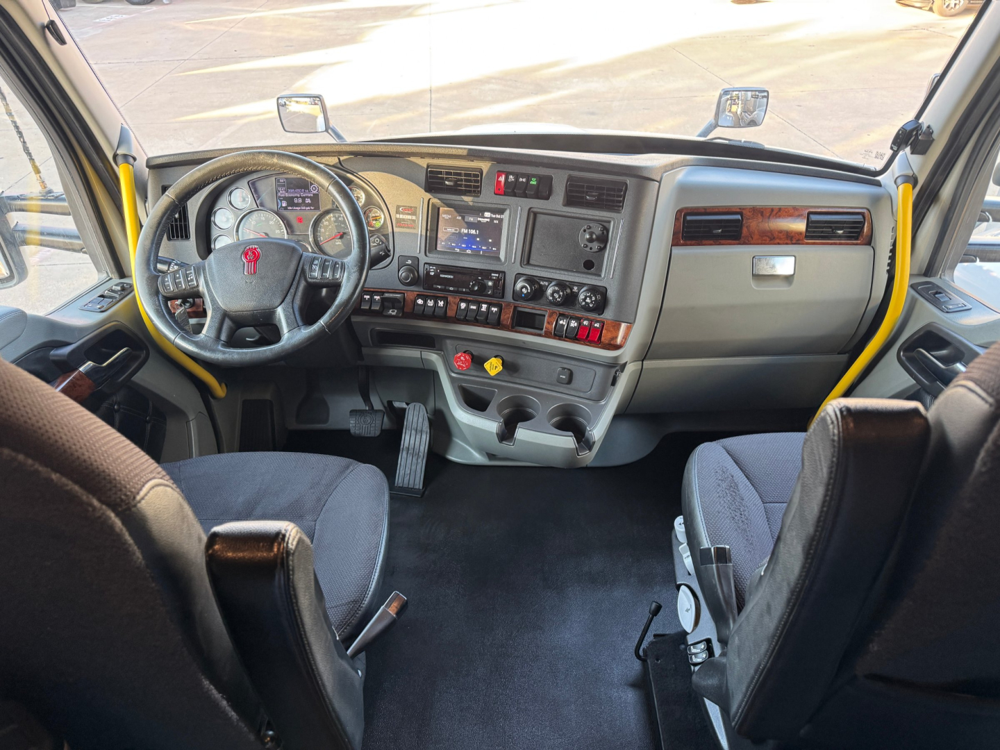 Kenworth truck cab interior dashboard with steering wheel, gauge cluster, and center console; yellow grab rails on both sides of the windshield; driver and passenger seats; various switches and controls.