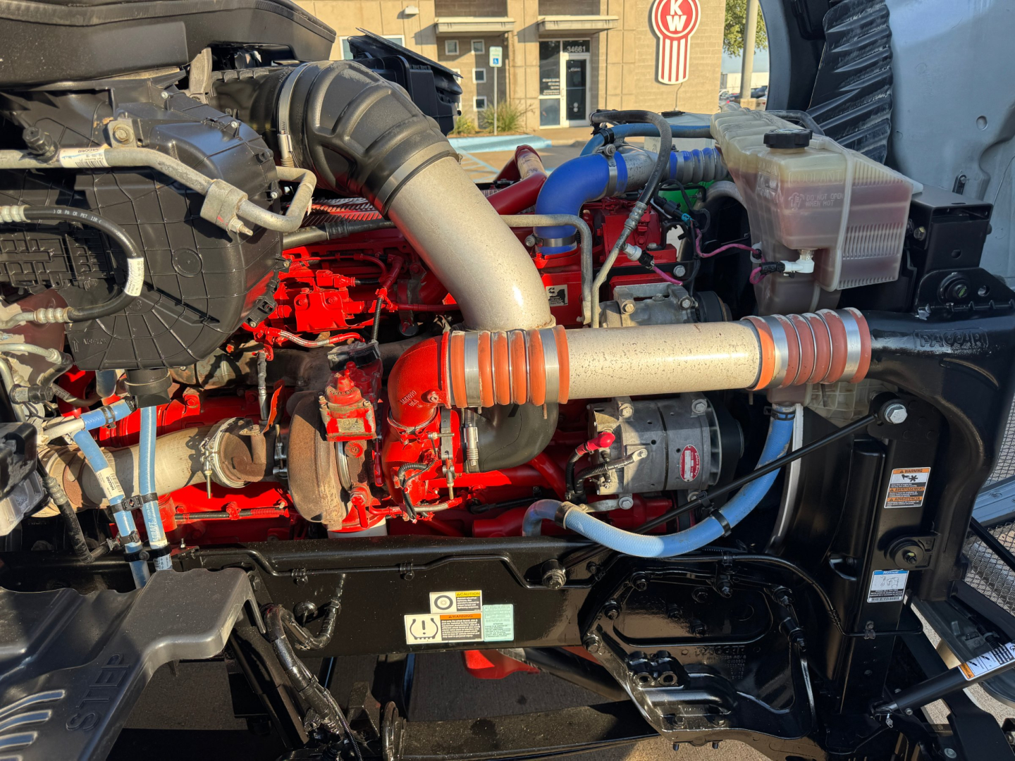 Kenworth T680 engine bay showing red engine block, intake tubing, and blue hoses with coolant reservoir.