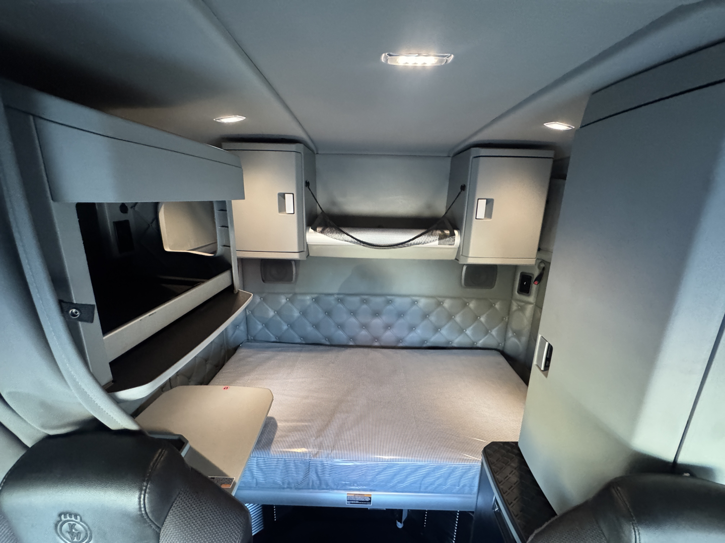 Kenworth T680 sleeper cab interior showing bed with quilted headboard, overhead cabinets, and side storage compartments.