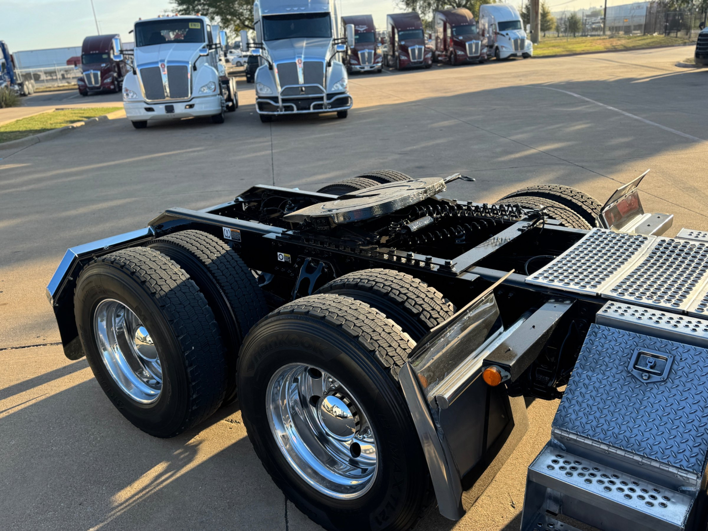 Kenworth truck chassis with dual rear axles and polished wheels; metal steps and fuel/tank area visible; multiple white trucks in background.