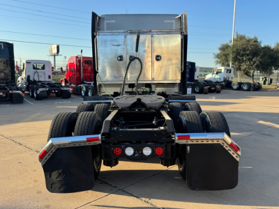 Kenworth T680 rear view showing fifth wheel, dual rear axles, mud flaps, and tail light cluster; chrome battery box and metal toolbox visible above; street lot with other trucks in background.