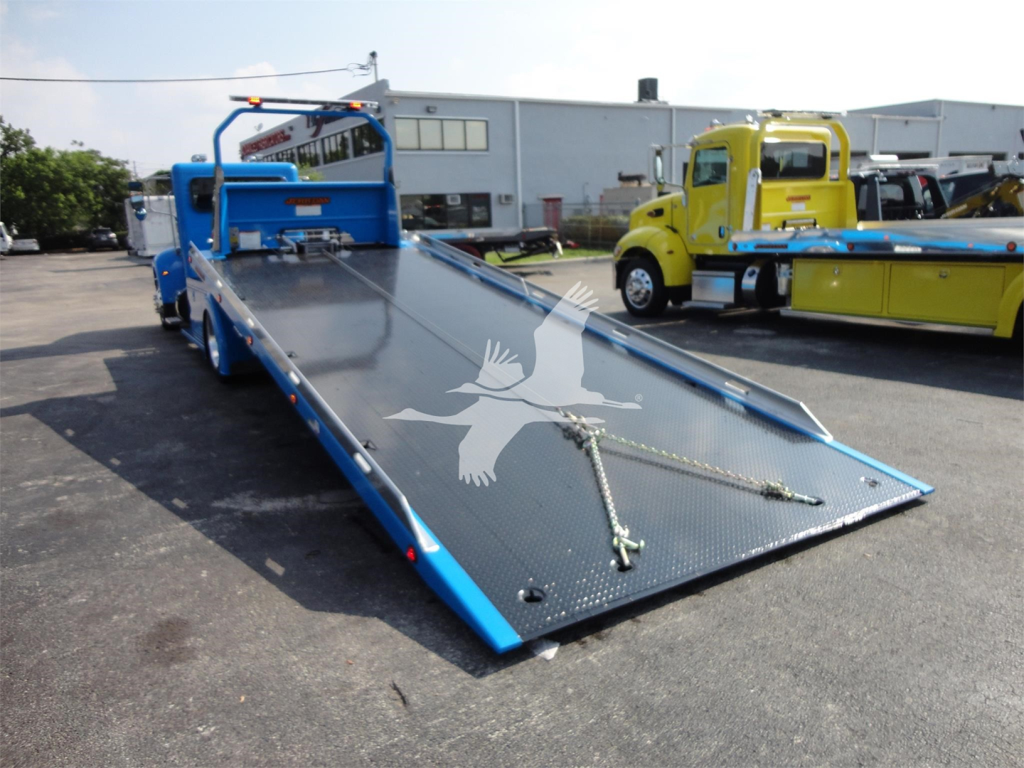 Blue Peterbilt 337 rollback tow truck flatbed with extended ramp in foreground, other colored trucks in background.
