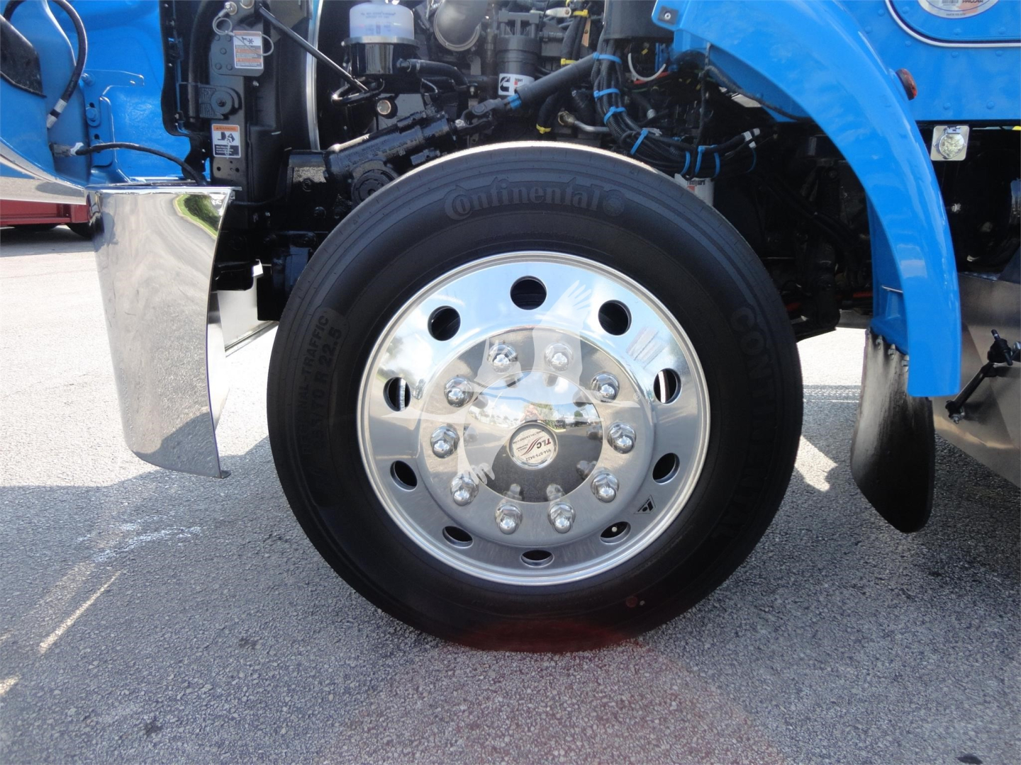 Front left wheel of a blue Peterbilt truck, chrome hub with multiple lug nuts, Continental tire, engine bay visible with various hoses and components.
