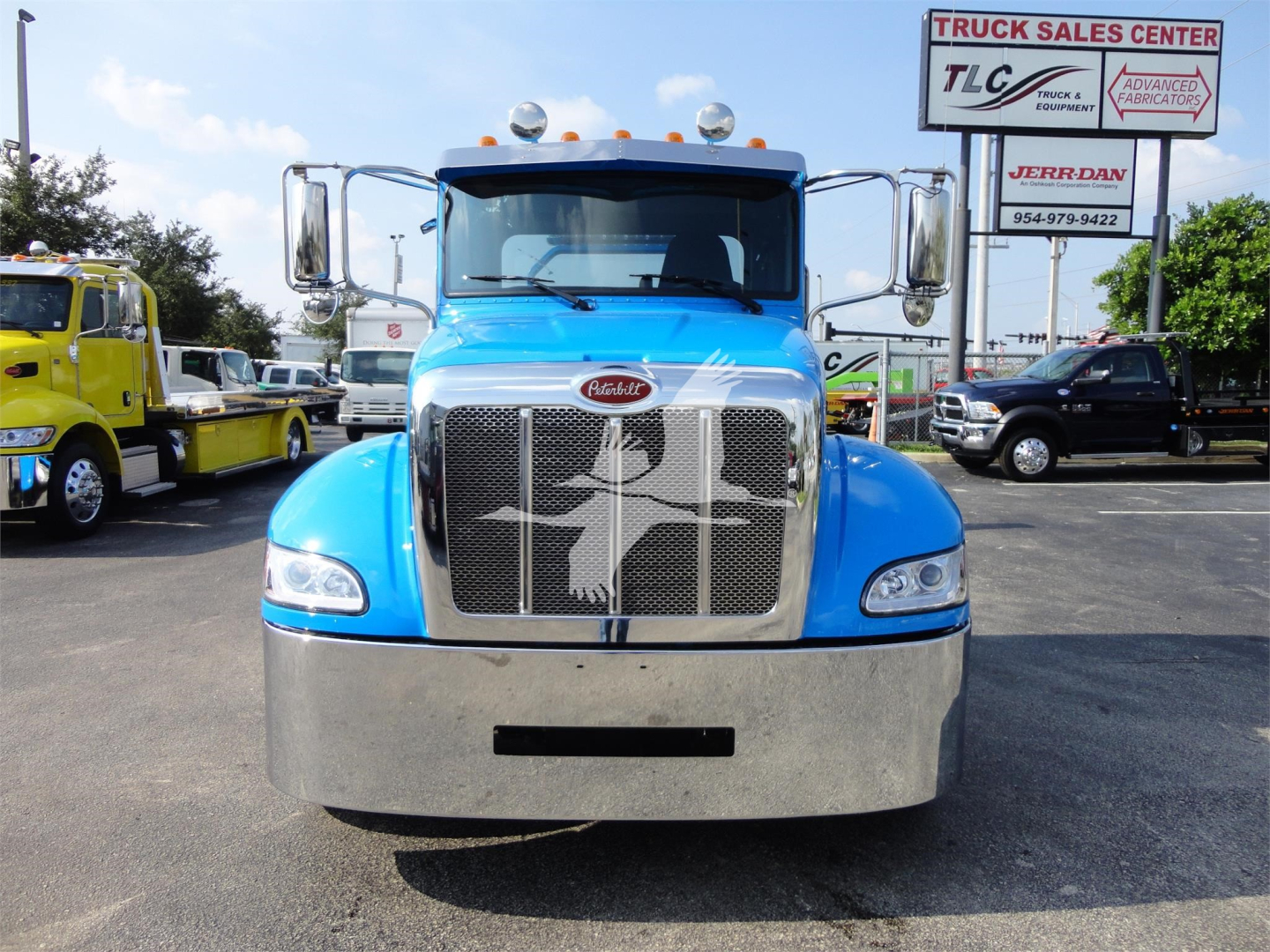 Front view of a blue Peterbilt 337 truck in a dealership lot with chrome grille, large bumper, and side mirrors; other trucks and signages visible in background.