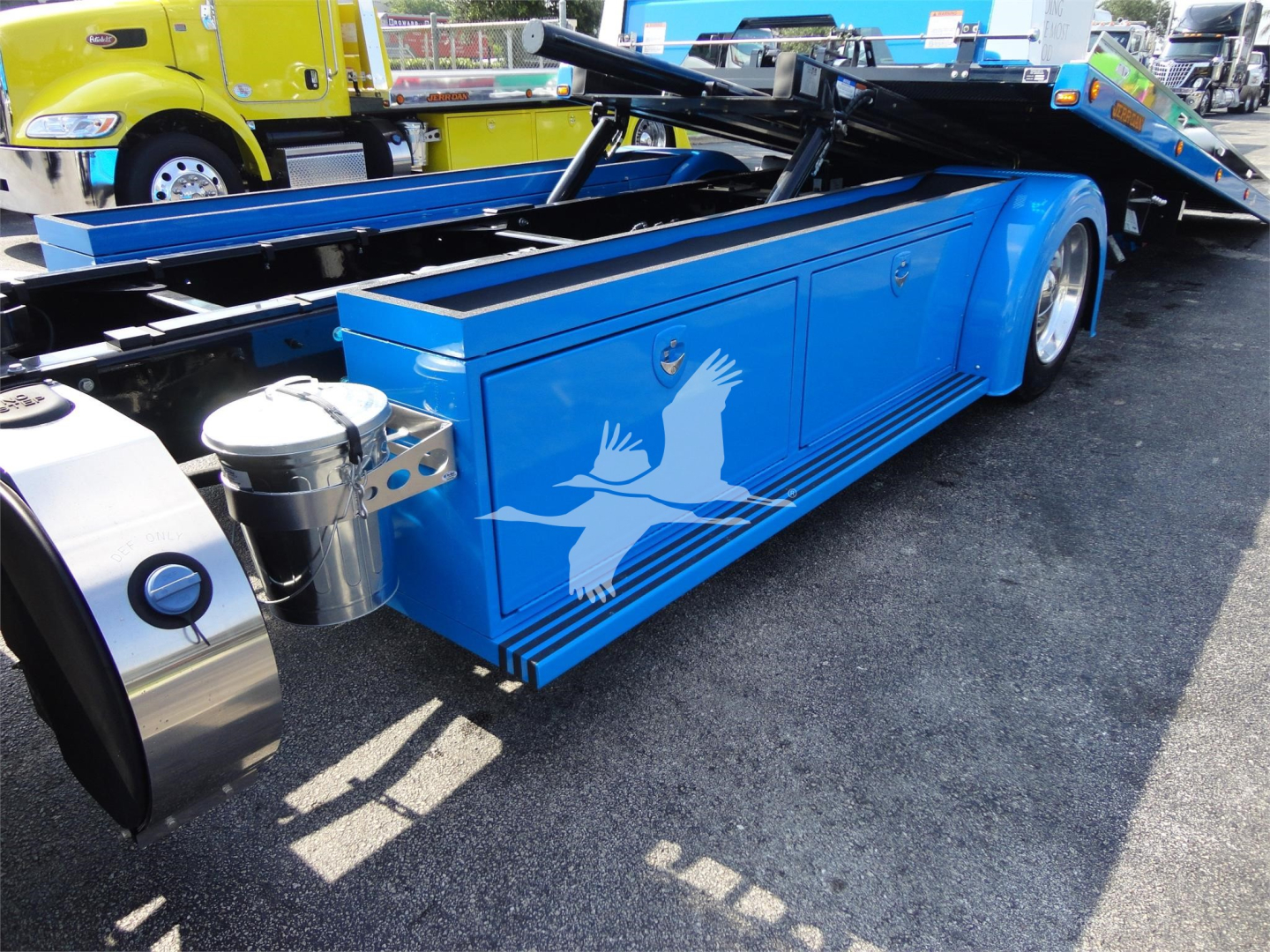 Truck body with blue tool/storage compartments and white bird graphic on side, Peterbilt 337 rollback in a dealership lot.